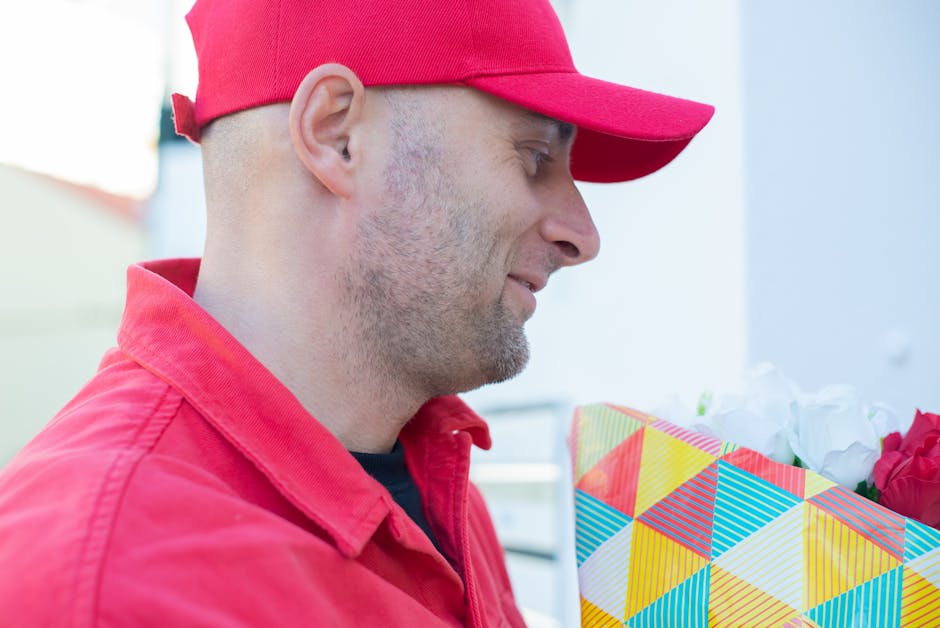A man wearing a red cap and matching red jacket holds a colorful, geometric-patterned gift bag filled with white and red flowers, ready for flower delivery. The arrangement features fresh petals and g