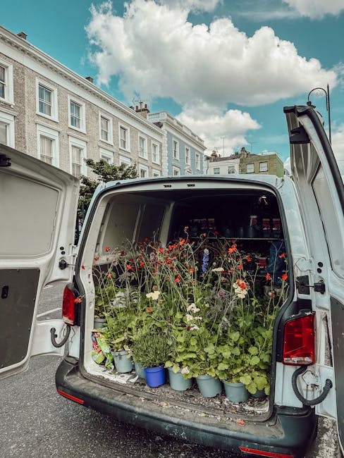 The open rear of a white delivery van filled with potted flowers, including red, white, and green foliage, parked on a city street with classic Georgian-style buildings and a partly cloudy sky overhea