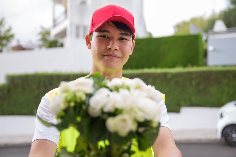 A delivery person wearing a red cap and a high-visibility vest is smiling warmly while presenting a bouquet of fresh white roses and green foliage. The bouquet is arranged in a compact, rounded style,